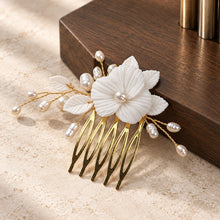 Charger l'image dans la galerie, Peigne cheveux mariage fleur blanche en porcelaine et perles naturelles, finition dorée, présenté devant un socle en bois foncé.