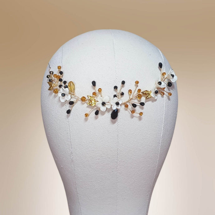 Accessoire de cheveux noir et doré aux fleurs blanches, présenté sur support avec fond beige.