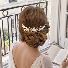Charger l'image dans la galerie, Mariée assise près d’une fenêtre portant un bijou floral en porcelaine blanche sur un chignon bas sophistiqué