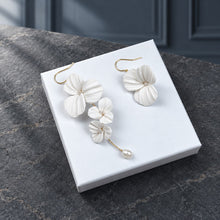 Charger l'image dans la galerie, Boucles d’oreilles asymétriques posées sur un présentoir blanc, avec fleurs blanches en porcelaine froide et perles.
