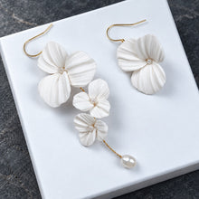 Charger l'image dans la galerie, Vue rapprochée des deux boucles d’oreilles asymétriques : fleurs blanches en porcelaine froide, perles et finitions dorées.