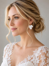 Charger l'image dans la galerie, Boucle d’oreille florale en porcelaine blanche et perles, portée par une mariée blonde avec chignon bas et robe de mariée en dentelle.