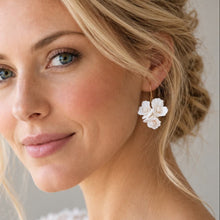 Charger l'image dans la galerie, Boucle d’oreille de mariée avec fleurs blanches en porcelaine froide et perles, portée avec un chignon blond et une robe en dentelle.