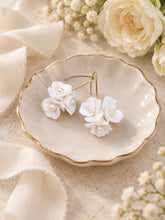 Charger l'image dans la galerie, Paire de boucles d’oreilles florales de mariage avec fleurs en porcelaine blanche, perles nacrées et crochets dorés, présentée dans une coupelle ivoire.
