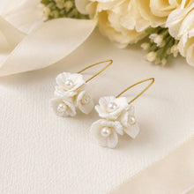 Charger l'image dans la galerie, Deux boucles d’oreilles de mariée avec fleurs blanches façonnées en porcelaine froide, perles et attaches dorées, posées sur un support clair.