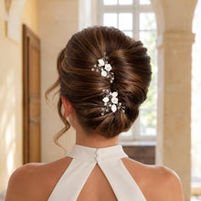 Charger l'image dans la galerie, Deux épingles de mariage à fleurs et feuilles blanches en porcelaine froide, perles naturelles et cristaux transparents, placées l’une sous l’autre dans un chignon banane brun