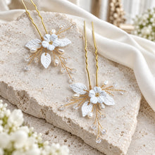 Charger l'image dans la galerie, Deux épingles aux finitions dorées, ornées de fleurs et feuilles blanches en porcelaine froide, de perles naturelles et de cristaux transparents présentées à plat sur pierre naturelle