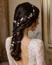 Charger l'image dans la galerie, Une longue vigne de cheveux flexible ornée de feuilles métalliques en or rose, de petites fleurs blanches et de minuscules perles transparentes. L'accessoire est mis comme couronne, puis tressé dans une coiffure brune, descendant jusqu'au bas de la tresse.
