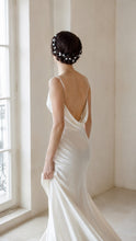 Charger l'image dans la galerie, Vigne de cheveux à feuilles blanches portée en couronne sur une tresse enroulée autour de la tête par une mariée brune en robe blanche dos nu.